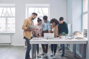 Group of happy young people in smart casual wear discussing business while standing near desk in the office photo