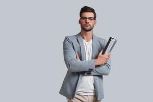 Thoughtful handsome. Good looking young man in eyeglasses keeping arms crossed and looking at camera while standing against grey background photo