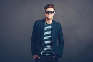 Look at me now. Confident young handsome man in sunglasses keeping hands in pockets and looking at camera while standing against grey background photo