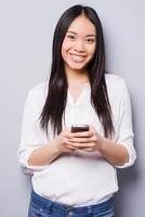 Beauty with phone. Cheerful young Asian woman holding mobile phone and smiling while standing against grey background photo