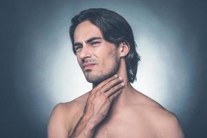 sintiendo dolor en la garganta. retrato de un joven sin camisa que expresa negatividad mientras toca su cuello y se enfrenta a un fondo gris foto