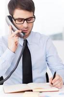 concentrado en el trabajo. un joven confiado con camisa y corbata hablando por teléfono móvil y escribiendo algo en un bloc de notas mientras se sienta en su lugar de trabajo foto