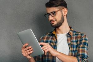 Enjoying his new digital tablet. Confident young man using his digital tablet and looking at it while standing against grey background photo