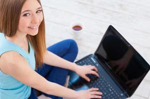 Spending time in the net. Top view of cheerful teenage girl using computer and smiling while sitting on the floor at her apartment photo