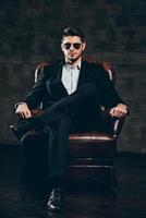 Elegance and masculinity.Young handsome man in suit and sunglasses holding hand on chin and looking at camera while sitting in leather chair against dark grey background photo