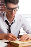 Confident and creative. Handsome young man in shirt and tie writing something in note pad while sitting at his working place photo