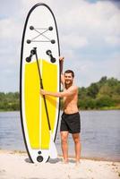 listo para remar. apuesto joven sosteniendo su paddleboard mientras camina por la orilla del río foto