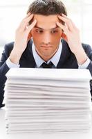 That is too much. Depressed young man in shirt and tie looking at the stack of paperwork and holding head in hands while sitting at his working place photo