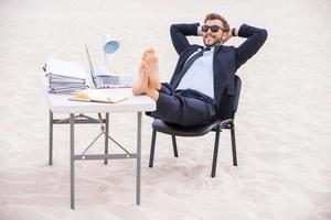 listo para vacaciones. un joven apuesto con ropa formal y gafas de sol cogidos de la mano detrás de la cabeza y sosteniendo los pies sobre la mesa de pie sobre la arena foto