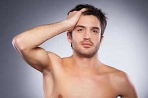 Just woke up. Handsome young beard man looking at camera and holding hand in hair while standing isolated on grey background photo