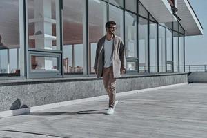 Modern businessman on the go.  Full length of confident young and handsome man smiling and looking away while walking outdoors photo