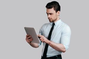 Examining his digital tablet.  Serious young man in white shirt and tie working on his digital tablet while standing against grey background photo