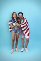 toda la longitud de una hermosa pareja joven cubierta con una bandera estadounidense sonriendo mientras se enfrenta a un fondo azul foto