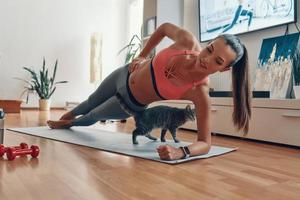 Attractive young woman in sports clothing standing in plank position with cat walking around while exercising at home photo