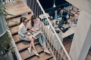 Top view of two young modern women in smart casual wear communicating and using laptop while sitting on the stairs photo