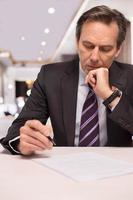 Signing a contract. Thoughtful mature man in formalwear signing a document and holding hand on chin photo