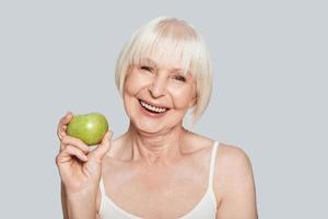 Full of vitamins. Beautiful senior woman holding an apple and smiling while standing against grey background photo