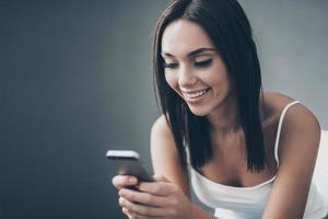 enviando mensajes de texto a un amigo. atractiva mujer joven sosteniendo un teléfono inteligente y mirándolo con una sonrisa mientras se sienta contra la pared gris foto