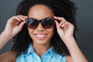 Ready to sunny days. Portrait of attractive young African woman adjusting her sunglasses and smiling while standing against grey background photo