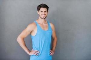 Fit and confident. Happy young muscular man holding hands on hip while standing against grey background photo