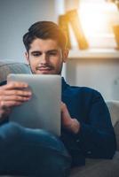 Enjoying his new digital tablet. Close-up of handsome young man using his digital tablet with smile while lying down on the couch at home photo