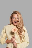 Surprised young smiling woman holding a flower and looking at camera while standing against grey background photo