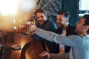 Happy young men in casual clothing toasting each other with beer and laughing while sitting in the pub photo
