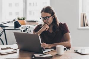 Solutions every day. Thoughtful young man with long hair looking at laptop and holding hand on chin while sitting at his working place in office photo