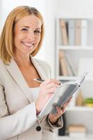 Taking notes with smile. Smiling mature businesswoman writing in note pad and looking at camera while leaning at the wall photo