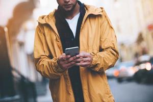 Quick message on the go. Close-up of handsome young man holding smart phone and looking at it while standing on the city street photo