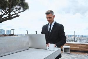 Confident mature man working on laptop while standing on the rooftop terrace photo