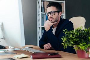 Thinking about solution. Thoughtful young man in smart casual wear holding hand on chin and looking at computer monitor while sitting at his working place in office photo