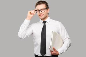 hombre de negocios con laptop. un joven apuesto con camisa y corbata ajustando sus gafas y sosteniendo una laptop mientras se enfrenta a un fondo gris foto