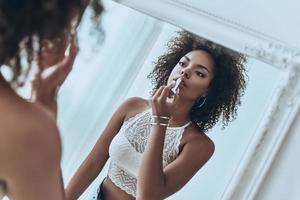 Adding some shine to her lips. Reflection of attractive young African woman putting on lipstick while standing in front of the mirror at home photo