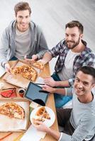 Tasty food for working good. Top view of three cheerful young men eating pizza while sitting at the desk together photo