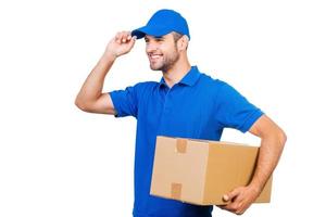 Delivering in pleasure. Happy young courier holding a cardboard box and adjusting his cap while standing against white background photo