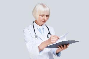 Taking care of paperwork. Beautiful senior woman in lab coat holding a clipboard with papers while standing against grey background photo