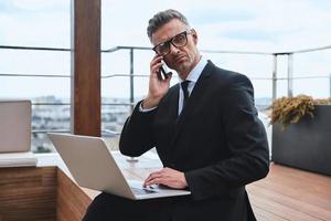 Confident mature man working on laptop and using mobile phone while standing on the rooftop terrace photo