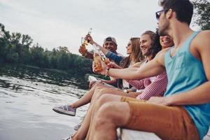 Relaxing among friends. Group of happy young people in casual wear smiling and drinking beer while sitting on the pier photo