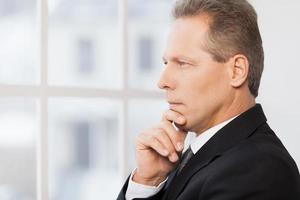 Thinking about his business. Side view of thoughtful mature man in formalwear holding hand on chin and looking away photo