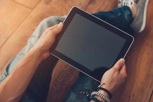 All the world in my hands. Close-up top view of man holding digital tablet while sitting on the wooden floor photo