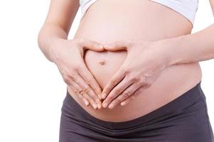 I love my baby. Cropped image of pregnant woman holding hands in heart shape on her abdomen while standing isolated on white photo