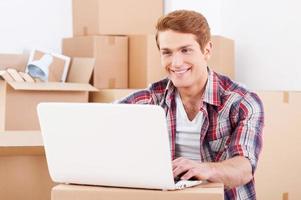 In search of a good moving company. Handsome young man sitting on the floor and working on laptop while cardboard boxes laying in the background photo