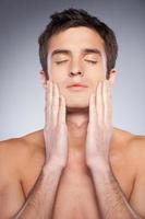 Smooth and healthy skin. Handsome young shirtless man touching his face with hands and keeping eyes closed while standing against grey background photo