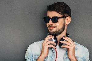 Music and style are his passion. Young man adjusting his headphones and looking away with smile while standing against grey background photo