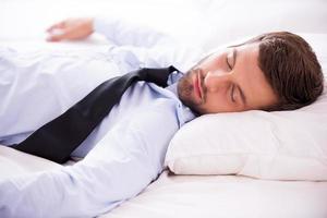 sentirse agotado después del día de trabajo. apuesto joven con camisa y corbata durmiendo en la cama foto