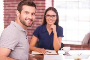 Job candidate. Handsome young man sitting at the table and looking over shoulder with smile while cheerful woman sitting in front of him photo