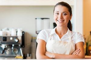 barista confiado. hermosa joven en delantal con los brazos cruzados mientras está de pie en la cafetería foto