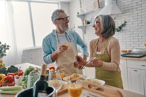 pareja senior moderna en delantales preparando una cena saludable y sonriendo mientras pasa tiempo en casa foto