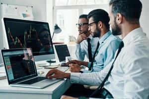 Working with analytics. Group of young modern men in formalwear analyzing stock market data while working in the office photo
